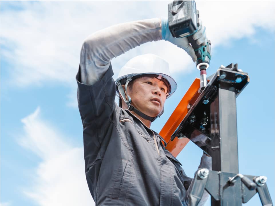 ヘルメットをかぶった作業員が、青空の下でドリルを使って金属構造を取り付けている。