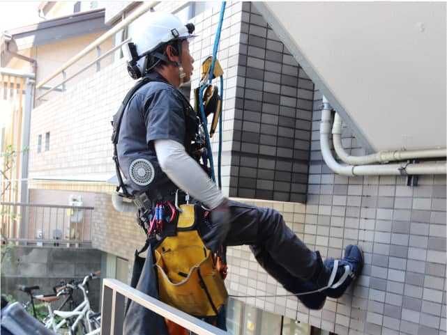 高所作業用の装備を身につけた作業員が、建物の壁で作業をしている。