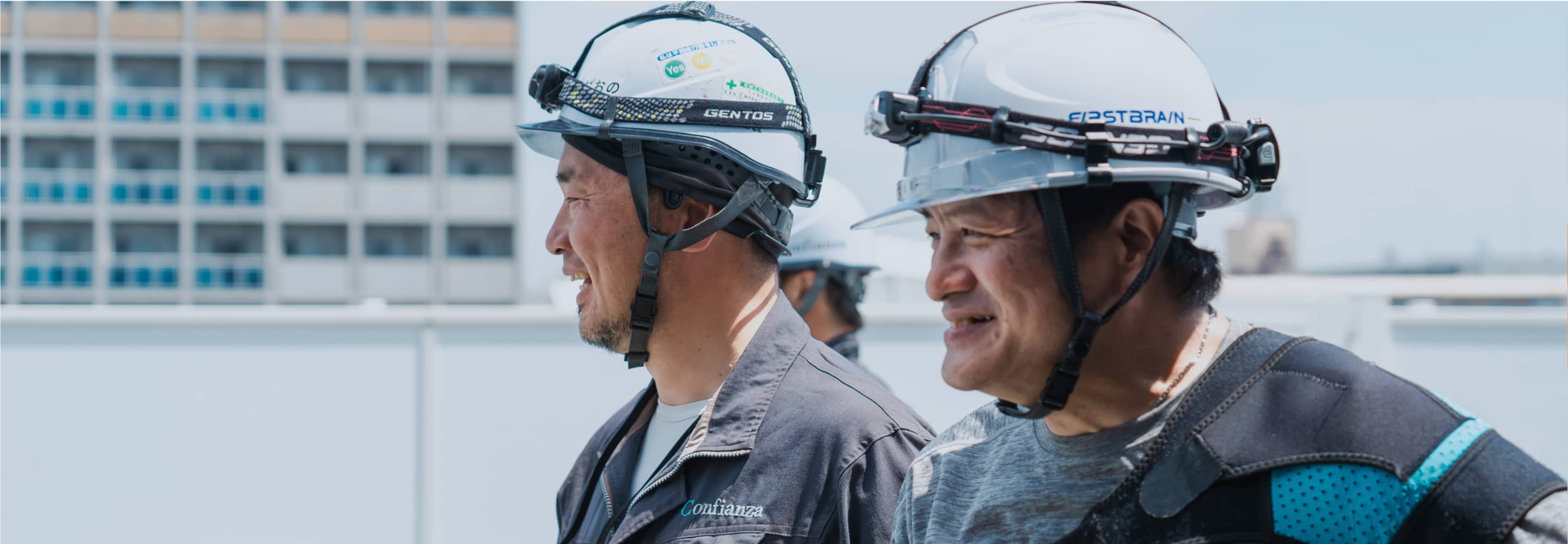 ヘルメットをかぶった2人の作業員が屋外で笑顔で話している。