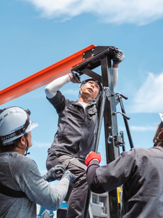 鉄骨を支える作業員が、青空の下で作業している。