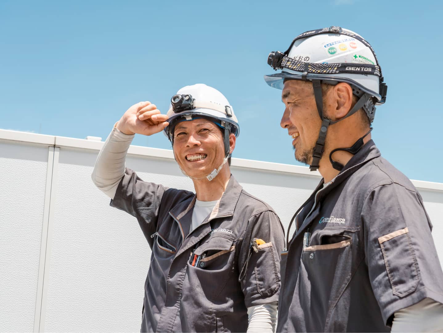 安全装備を着用した笑顔の作業員たちが青空の下で話している。