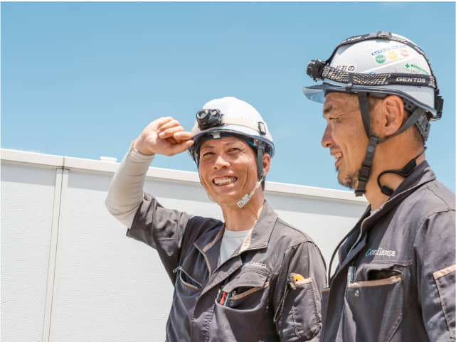 ヘルメットをかぶった作業員が屋外で笑顔で会話している。
